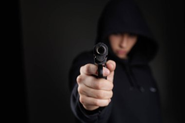 İsimsiz kapüşonlu kişi, silah tehdidi, şiddet riski ve karanlık arka planda kamu güvenliği veya güvenlik kavramını gösteren izleyiciye silah doğrultuyor
