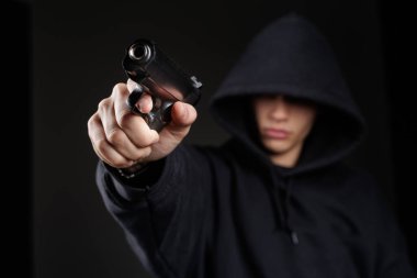 Kapüşonlu kişi, silahlı soygun tehdidi ve güvenlik riski kavramını temsil eden karanlık bir sahnede kameraya silah doğrultuyor.