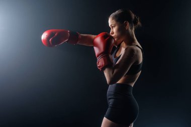 Kırmızı eldivenli kadın boksörün karanlık stüdyo arkaplanını web sitesi afişi ya da sosyal reklam için fotokopi alanı ile yumrukladığı yan görüntü.