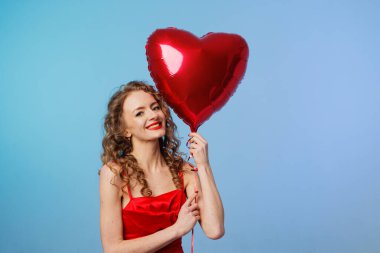 Kırmızı elbiseli gülümseyen kadının stüdyo portresi mavi arkaplanlı kalp balonu taşıyor. Fotokopi alanı var, afiş veya sosyal sevgililer günü kampanyası için.
