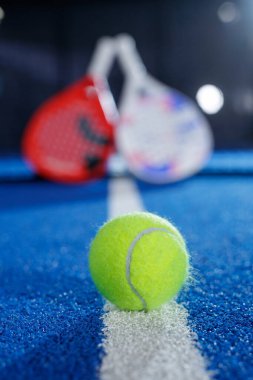 Mavi bir kortta, arkasında odağını kaybetmiş raketlerle bir Padel tenis topunun yakın çekimi. Reklam, web sitesi afişleri, spor tanıtımı ve görsel markalaşma için ideal.