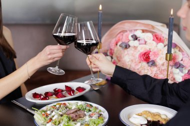 Mum ışığında, çiçeklerle ve gurme yemeklerle dolu samimi bir akşam yemeğinde kırmızı şarap kadehleri tokuşturan çift. Konukseverlik, flört ve görsel pazarlama için ideal..