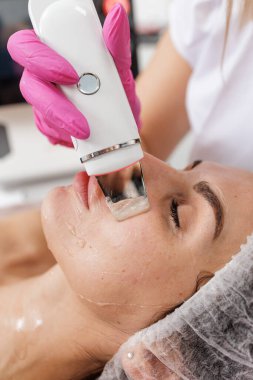 Kozmetik uzmanı kadın hastaya deri temizleme cihazı kullanarak ultrasonik iyon temizleme yapıyor. Dermatoloji, estetik cilt bakımı ve profesyonel güzellik servisi tanıtımı için ideal..