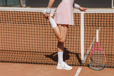 Dişi tenisçi dışarıda raket tutarak bacak bacak esnetiyor. Spor reklamcılığı, fitness kampanyaları ve yaşam tarzı promosyonu için ideal..