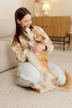 Mutlu kadın halıda oturup Corgi köpeğine sarılıyor. Ev sahibi ile evcil hayvan arasındaki sevgi, arkadaşlık ve duygusal bağı gösteren sıcak bir ev hayatı..