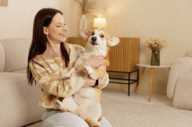Sıcak oturma odasında yüzünde komik bir ifade olan sevimli bir Corgi tutan gülümseyen kadın. Evcil hayvan sahipliği, arkadaşlık ve neşeli ev hayatı kavramı..