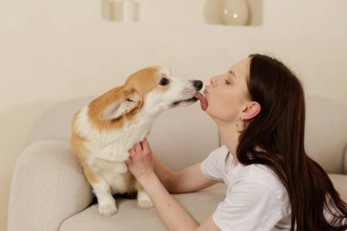 Kanepede oturan genç bir kadın köpek yüzünü yalarken usulca korgisini tutuyor. Sevgi, güven ve neşe dolu dostluk gösteren sıcak bir aile anı..