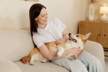 Kanepede rahat bir şekilde Corgi köpeğini kucağında tutan gülümseyen kadın. Evcil hayvan ve ev sahibi arasındaki sevgi, güven ve neşeli arkadaşlığı gösteren kapalı bir yaşam tarzı..