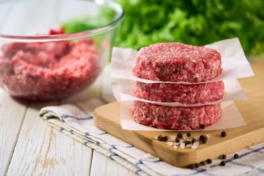 Beyaz ahşap bir masa üzerinde parşömen kağıdıyla ayrılmış çiğ hamburger köfteleri yığını. .