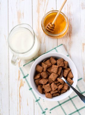 Süt ve bal dolu bir kasede çikolatalı mısır pedleri beyaz ahşap bir masada..