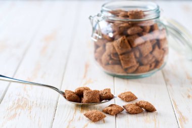 Hafif mutfak masasındaki metal kaşıkta çikolatalı mısır pedleri seçici bir odak noktası. Cam kavanozda tahıl gevreği ve ahşap masaya dökülüyor..