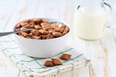 Beyaz ahşap bir masada seramik kasede çikolatalı mısır pedleri, seçici bir odak noktası. Mısır yastığı ve süt kullanarak sağlıklı bir kahvaltı gevreği hazırlıyorum..