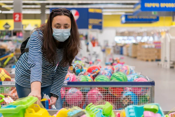 A mulher em uma máscara protetora em um supermercado escolhe brinquedos de crianças. Mulher em máscara médica no supermercado tonificado — Fotografia de Stock
