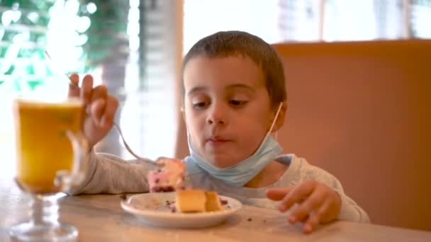 Un ragazzino con una maschera medica in un bar mangia una torta. Un bambino che indossa una maschera in un caffè durante una pandemia — Video Stock