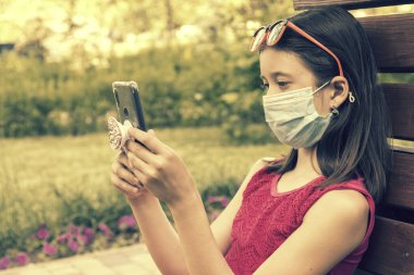 Akıllı telefon tutan koruyucu maskeli bir kız. Green Park 'taki bankta oturan genç bir kız. Cep telefonuyla. Koronavirüs kısıtlamalarının gevşetilmesi ve kendini izole etmenin ilk aşaması. ses tonlu