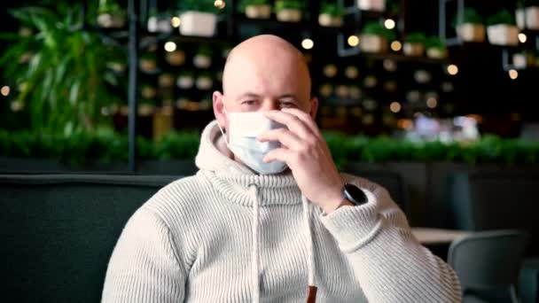 El hombre se quitó la máscara para beber capuchino en la terraza. Hombre en la cafetería al aire libre durante la cuarentena. Covid-19 Concepto de desayuno y pausa para café. Calvo chico en un café bebidas capuchino — Vídeos de Stock