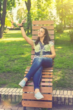 Parktaki bankta oturmuş, alışveriş torbalarıyla alışveriş yaptıktan sonra neşeli kız akıllı telefonuyla selfie çekiyor. Genç bir kadın selfie çekiyor, şehir parkında bir bankta oturuyor. Ton gibi. dikey resim