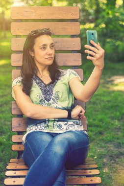 Parktaki bankta oturmuş, alışveriş torbalarıyla alışveriş yaptıktan sonra neşeli kız akıllı telefonuyla selfie çekiyor. Genç bir kadın selfie çekiyor, şehir parkında bir bankta oturuyor. Ton gibi. dikey resim