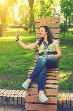 Parktaki bankta oturmuş, alışveriş torbalarıyla alışveriş yaptıktan sonra neşeli kız akıllı telefonuyla selfie çekiyor. Genç bir kadın selfie çekiyor, şehir parkında bir bankta oturuyor. Ton gibi. dikey resim