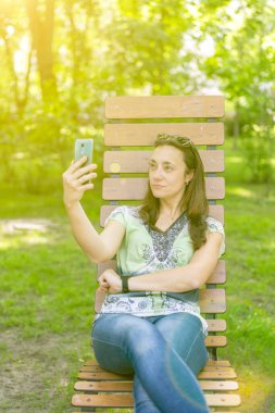 Genç bir kadın selfie çekiyor, şehir parkında bir bankta oturuyor. Ton gibi. dikey resim