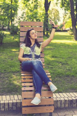 Parktaki bankta oturmuş, alışveriş torbalarıyla alışveriş yaptıktan sonra neşeli kız akıllı telefonuyla selfie çekiyor. Genç bir kadın selfie çekiyor, şehir parkında bir bankta oturuyor. Ton gibi. dikey resim