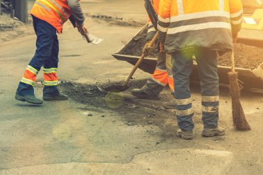 Parlak turuncu yansıtıcı üniformalı yol işçileri birikmiş kumu kazmak için kürek kullanırlar. Yol ve otoban kaldırımlarının bakımı. Çukur onarımı işe yarıyor. ses tonlu