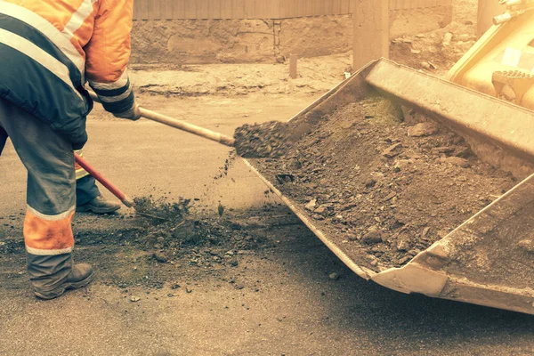 Los trabajadores del tráfico en uniformes reflectantes naranja brillante raspan arena acumulada y escombros entre carriles con palas y la cargan en un cubo de nivelador de metal. tonificado. — Foto de Stock