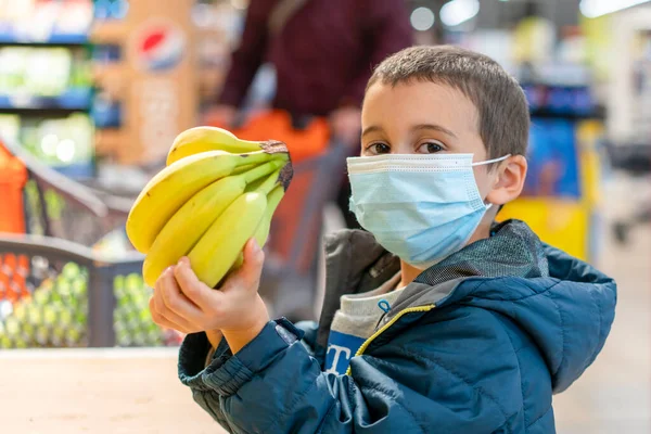 Egy gyerek, aki sebészeti maszkot visel, gyümölcsöt vásárol a szupermarketben a koronavírus világjárványban. Egy kisfiú egy szupermarketben orvosi maszkot visel. Coronavirus karantén. tónusú — Stock Fotó