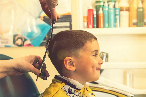 Little boy having a haircut at hair salon..Children hairdresser with scissors and comb is cutting little boy. Contented cute preschooler boy getting the haircut. toned — Stock Photo, Image