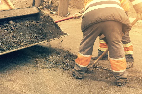 Los trabajadores del camino en uniformes reflectantes naranja brillante utilizan palas para raspar la arena acumulada. mantenimiento de pavimentos de carreteras y carreteras. trabajos de reparación de baches. tonificado — Foto de Stock