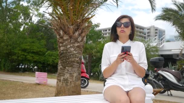 La ragazza è seduta su una panchina e usa il telefono — Video Stock