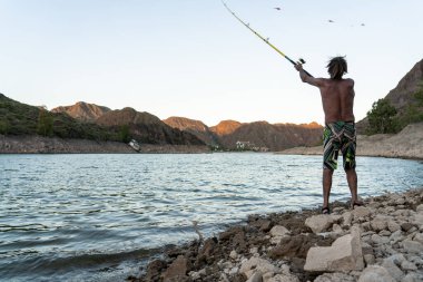 Sakin su ve günbatımı dağlarıyla kayalık göl kıyısında balık tutan bir adamın arka görüntüsü..