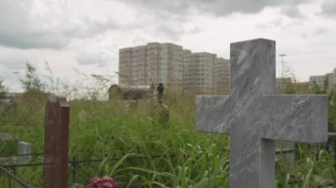 Terk edilmiş bir mezarlıkta gri mermer bir haç. Apartman evinin arka planında ve yeşil, uzun çimen tarlasında. Mezar dekorasyonu. Sevdiklerini ezberlemek. Bulutlu gökyüzü. Eski mezarlık.