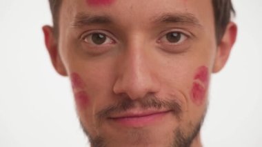 Gorgeous man with light in brown eyes, moustache, red kiss marks on face looking straight, smiling on white wall background close up. Portrait one handsome male in love with stains of lip gloss stick.