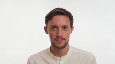 Beyaz, kendine güvenen, kısa, koyu kahverengi saçlı, sakallı, bıyıklı, beyaz arka planı yakından düşünen genç beyaz bir adam. Yüzünde öpücük izi olan adamın portresi.