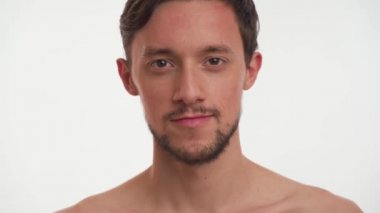 Koyu renk saçlı, yarı çıplak sakallı yakışıklı adam elinde pembe ruj tutuyor, açıyor, gülümsüyor, flört ediyor, beyaz arka planda izole edilmiş kameraya bakıyor. Seksi moda erkeğinin yakın portresi..