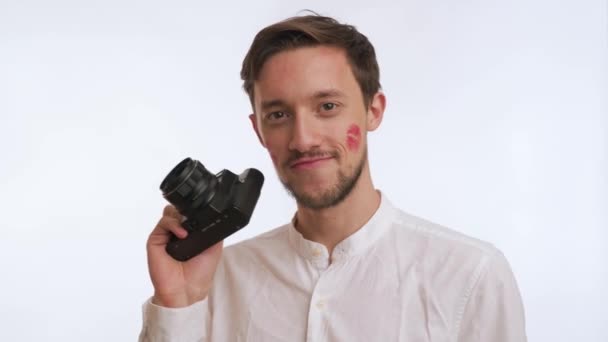 有胡子的美国男人的肖像 红唇吻印或脸颊上的痕迹 穿着衬衫微笑 手持稀有的摄影相机 近距离拍摄白色背景 一个英俊的摄影师 — 图库视频影像