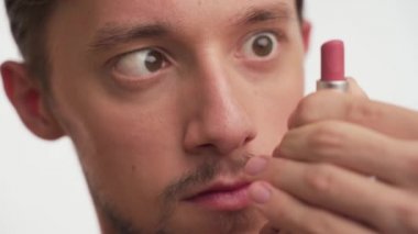 Close up portrait of one young stupid guy with brown eyes, hair, beard stare at pink lipstick, play with it, open and close on white background. Dumb male cross eyed, focus on women's accessory indoor