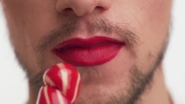 Close up mouth of gorgeous guy with make-up  red lipstick on lips. One young male with brown beard, moustache licking and sucking sweet sugar lollipop isolated on white background. Mouth view of man