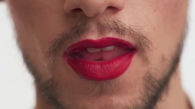 Dudaklarında ruj izi olan korkmuş esmer bir adam beyaz arka planda izole edilmiş kırmızı lolipopu yalıyor. Siyah sakallı, şeker bağımlısı genç bir adamın yüz ifadesi..