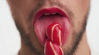 Makyajlı, kırmızı rujlu, dudakları açık, ağzı açık, diliyle şeker lolipopu yalayan yakışıklı bir adam. Siyah sakallı, beyaz arka planda izole edilmiş bıyıklı, şehvetli genç bir erkek. Erkek portresi.