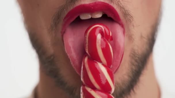 ホットハンサムなひげを生やした男は 唇に赤い口紅を着用し 彼の前に甘い砂糖菓子を保持し クリックして白い背景のクローズアップで喜びでそれを吸う 女性の裏切りのヒント 正面図 — ストック動画
