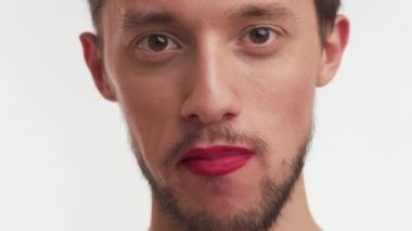 Sakallı, bıyıklı, kendine güvenen yakışıklı bir adam, makyajlı kırmızı ruj sürüyor, kameraya yakından bakıyor, lezzetli şekerlemeler yiyor, ısırıyor, beyaz arka planda izole edilmiş bir şekilde çiğniyor..