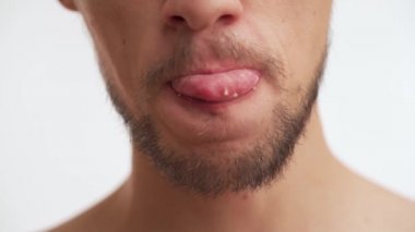 Koyu kahverengi sakallı, bıyıklı, dudak yalayan, yemek çiğneyen, ağır çekimde yutan genç beyaz bir adam. Beyaz duvar arka planında izole edilmiş tek bir erkek ağzı görüntüsü.