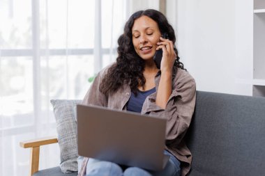 Kadın akıllı telefonda konuşuyor ve evdeki koltukta otururken dizüstü bilgisayar kullanıyor.