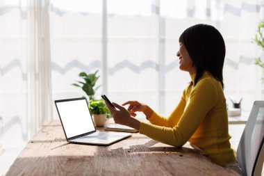 Ahşap bir masada akıllı telefon ve laptop kullanan bir kadın gülümsüyor.