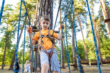 Yazlık giysili, emniyet kemeri ve ipe bağlı miğferi olan sevimli çocuk yeşil ağaçlar üzerinde yavaşça hareket ediyor. Güneşli bokeh arka planı. Spor, oyun, eğlence konsepti.