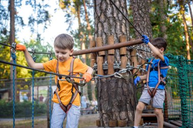 İki sevimli mutlu genç çocuk, çocuk ve kardeşi koruyucu koşum takımı içinde, karabina ve emniyet kaskları halatta, parlak güneşli bir günde yeşil yapraklı bokeh arka planda. Açık hava aktiviteleri, oyun konsepti.