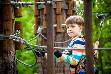Yazlık giysili, emniyet kemeri ve ipe bağlı miğferi olan sevimli çocuk yeşil ağaçlar üzerinde yavaşça hareket ediyor. Güneşli bokeh arka planı. Spor, oyun, eğlence konsepti.