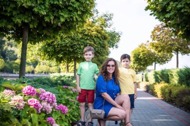 Genç bir kadın çocuklarıyla parkta oynarken eğleniyor. İlkokul çağındaki çocuklar. Aile pitoresk bitkilerle çevrili. Açık hava oyunları herkes için kullanışlı.